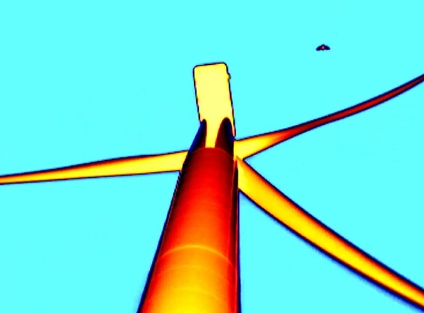 Thermal video still of bat approaching from downwind. The bat is black, the turbine is red-orange, and the sky is blue.