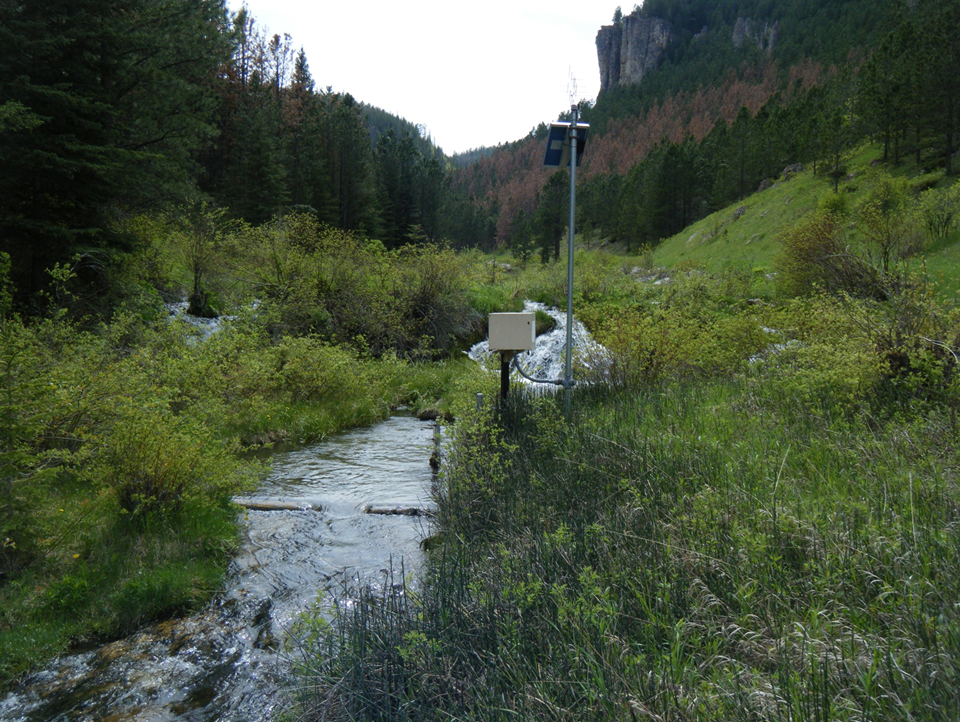 One of the types of sensors being used to capture the data. This photo shows a streamflow gage that measures water level or stage. Stage is then converted to discharge using a stage-discharge rating curve. On the pole is a solar panel that charges the battery. Above the panel is the transmitting antenna which communicates with the satellite to send data to the Internet in real time. The red trees on the hillside behind the gage are ponderosa pine trees recently killed by mountain pine beetles. If enough trees die, the water yield will increase streamflow at the gage.